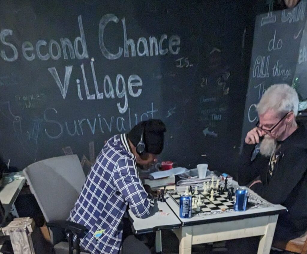 Two members play chess at a table in front of a chalkboard wall reading Second Chance Village