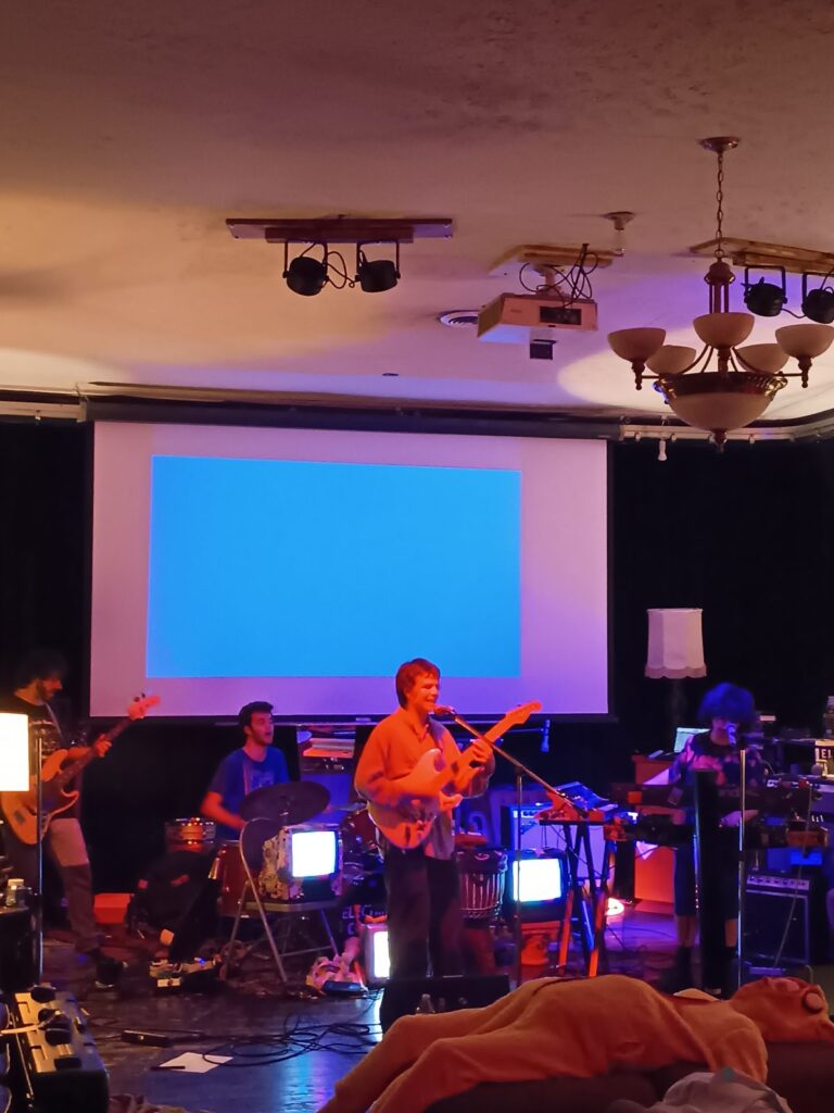 Wide view of a Second Chance Village live music night, with a band playing in front of a projection screen