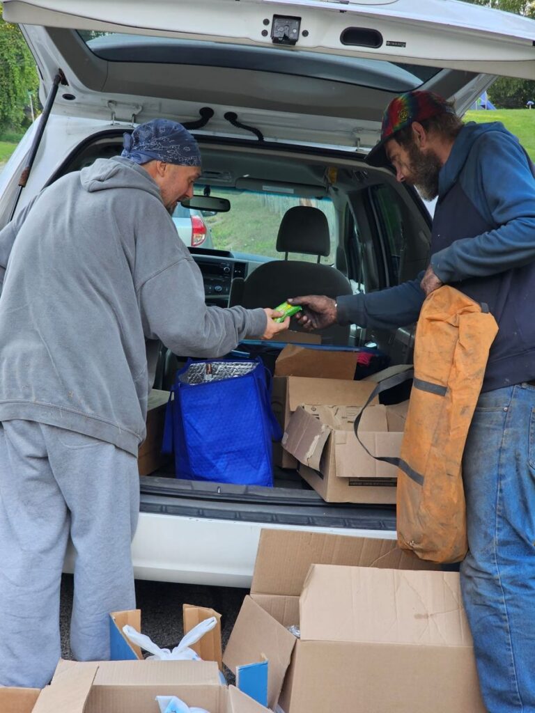 A Second Chance Village volunteer hands supplies to a community member from a car trunk full of boxes and bags