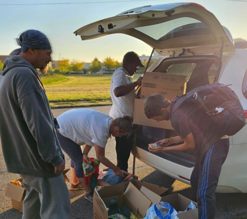 Second Chance Village volunteers hand out boxes of supplies from the back of an SUV at a park in the evening