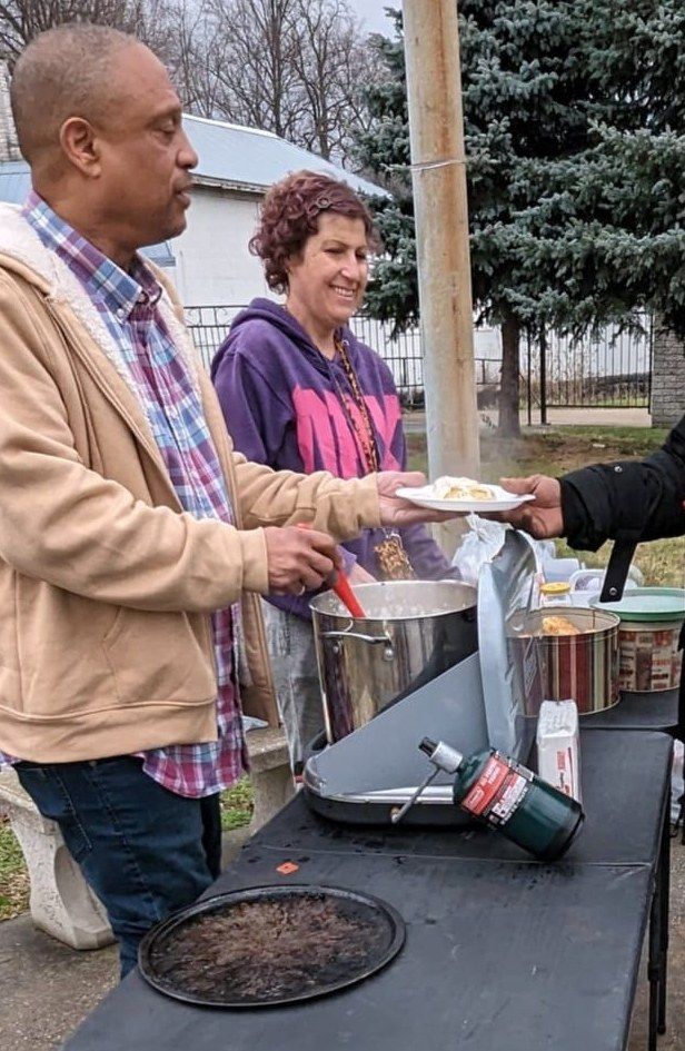 A Second Chance Village volunteer serves a plate of hot food from a large pot at an outdoor table